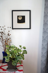 Corner of a room with a table, with an embroidered cloth, houseplant, and vase of dried flowers. A framed ink drawing of a moon over a barren landscape on a black background hangs on the wall.