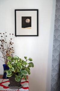 Corner of a room with a table, with an embroidered cloth, houseplant, and vase of dried flowers. A framed ink drawing of a moon over a barren landscape on a black background hangs on the wall.