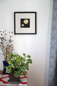 Corner of a room with a table, with an embroidered cloth, houseplant, and vase of dried flowers. A framed ink drawing of two moons on a black background hangs on the wall.
