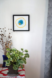 Corner of a room with a table, on which a houseplant and a vase of dried flowers are placed. A gray curtain hangs on one side. On the wall is a framed blue and yellow mixed media collage.