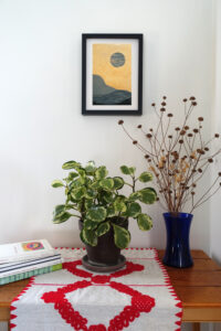 Corner of a room with a table. On the table are a pile of art books, a houseplant, and a vase of dried flowers. On the wall behind the table is a framed woodcut print of an abstracted landscape with a moon on light orange paper.