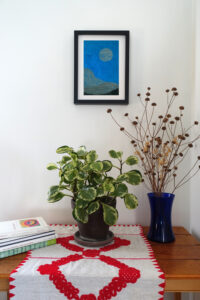 Corner of a room with a table. On the table are a pile of art books, a houseplant, and a vase of dried flowers. On the wall behind the table is a framed woodcut print of an abstracted landscape with a moon on blue paper.