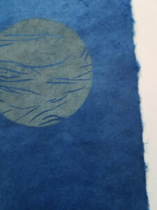 Detail of a woodcut print of an abstracted landscape with a moon above. The print is in shades of gray on dark blue paper.