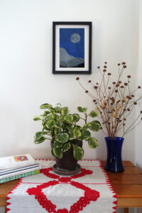 Corner of a room with a table. On the table are a pile of art books, a houseplant, and a vase of dried flowers. On the wall behind the table is a framed woodcut print of an abstracted landscape with a moon on navy blue paper.