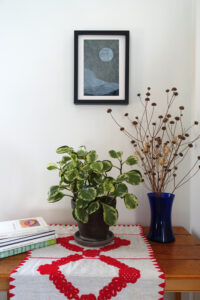 Corner of a room with a table. On the table are a pile of art books, a houseplant, and a vase of dried flowers. On the wall behind the table is a framed woodcut print of an abstracted landscape with a moon on gray paper.