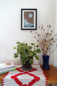 Corner of a room with a table. On the table are a pile of art books, a houseplant, and a vase of dried flowers. On the wall behind the table is a framed woodcut print of an abstracted landscape with a moon on brown paper.