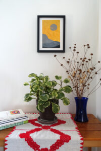 Corner of a room with a table. On the table are a pile of art books, a houseplant, and a vase of dried flowers. On the wall behind the table is a framed woodcut print of an abstracted landscape with a moon on orange paper.
