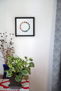 Corner of a room with a table, on which a houseplant and a vase of dried flowers are placed. A gray curtain hangs on one side. On the wall is a framed mixed media collage of small painted circles forming a ring.