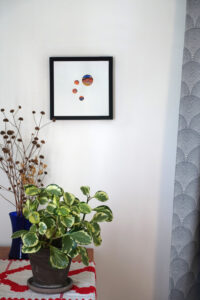 Corner of a room with a table, on which a houseplant and a vase of dried flowers are placed. A gray curtain hangs on one side. On the wall is a framed mixed media collage of painted blue and orange circles.