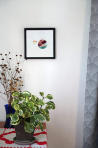 Corner of a room with a table, on which a houseplant and a vase of dried flowers are placed. A gray curtain hangs on one side. On the wall is a framed mixed media collage of two painted circles.