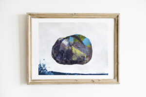 Mixed media painting of a single rock surrounded by white. The bottom edge of the paper is bordered by blue paint, which appears to splash upward on the left side. The work is on paper and is in a light wooden frame.